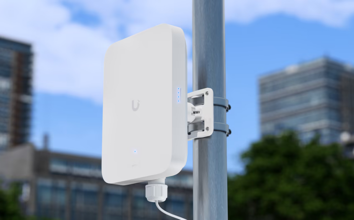 Ubiquiti U5G-Max-Outdoor UniFi 5G Max Outdoor installed on an outdoor pole with illuminated signal LEDs and a connected cable, with city buildings behind.