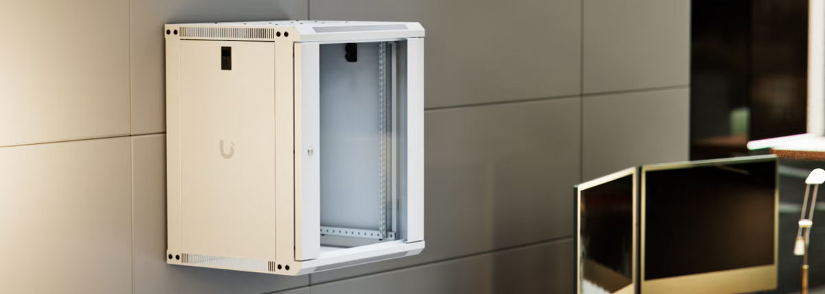 Ubiquiti UACC-Rack-12U-Wall-450-G UniFi Slim 12U rack cabinet wall-mounted above an office workstation with glass front door and internal rails visible.